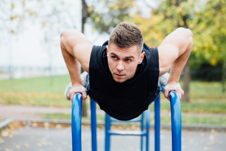 A portrait of a focused muscular young man in black workout clothes doing dips on parallel bars at...