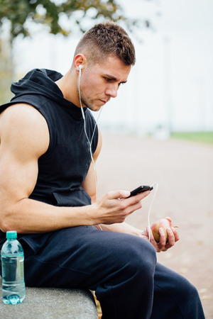 Fitness man wearing sportswear listening to music sitting on a stone bench. Relax after a hard workout, talking. White headphones.の写真素材