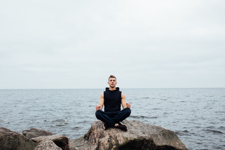 Strong fitness yoga man in lotus pose on the rock beach near the ocean. Harmonic concept, peace and successの写真素材