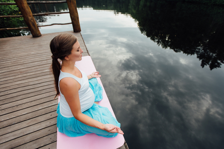 Young woman in the lotus position is practicing yoga in the forest next to the river. sitting on mats  the wooden pier.の写真素材