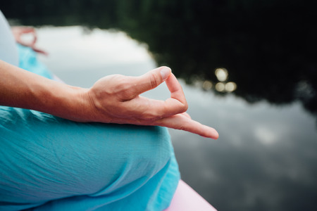 Woman meditating in the lotus position closeup. Hands close-up mudra. Sitting on wooden floor by the riverの写真素材
