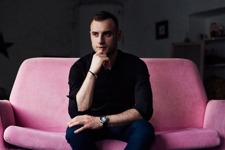 Man model sits on pink sofa in loft interior. Businessman in a black shirt and watch, blue pants.の写真素材