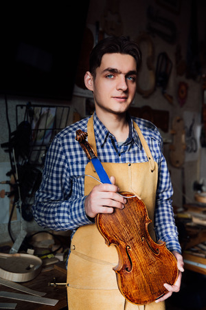 portrait of a young luthier with a handcrafted violin in his workshop with the tools.の写真素材