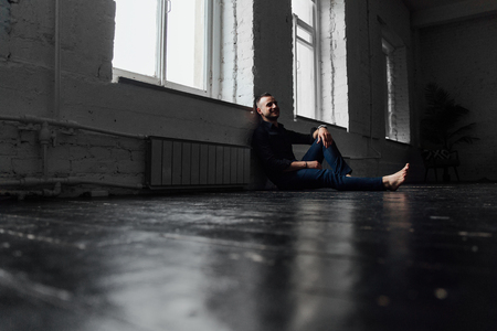 Portrait handsome fashionable man in a black shirt sits on a wooden floor by the window.の写真素材