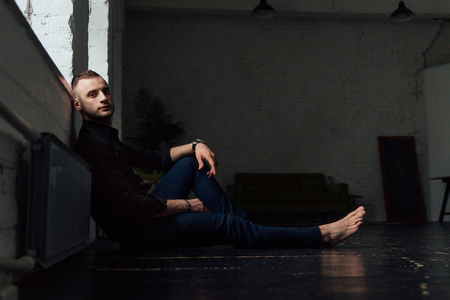 Portrait handsome fashionable man in a black shirt sits on a wooden floor by the window.の写真素材