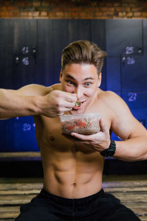 Fitness model of a bodybuilder guy eating food from a plastic container in a male locker room.の写真素材
