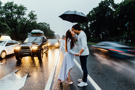The couple embraced under an umbrella rain, stands on the road. Cars are passing by.の写真素材