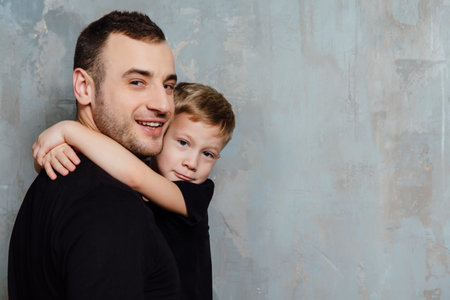 Father and son embracing son on gray wall background. Fashion man and boy in black clothes.