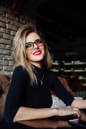 Young smiling business woman sitting in cafe at table, leaning hand on table and holding smartphone.の写真素材