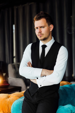 Stylish athletic man in a business costume vest. Sits in the turquoise sofa chair, a serious look.の写真素材