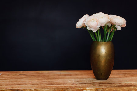 bouquet of pink delicate buttercups of flowers stands on a wooden table in a golden vase.