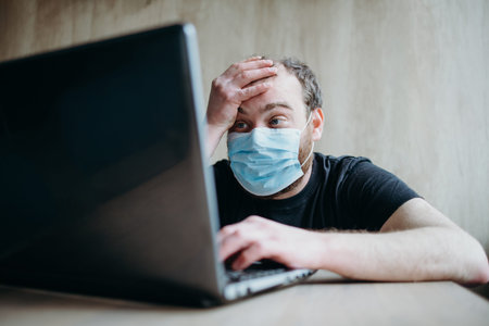 Coronavirus, an adult male with a beard works at a laptop. Protective respiratory mask.