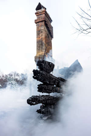 Remains of a house after a fire. The brick chimney of the stove and the charred planks are shrouded in dense smoke. Vertical background with a view of gloomy ash