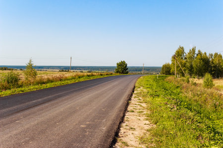 Freshly paved country road meanders past lush fields under a bright sky, offering a peaceful,...