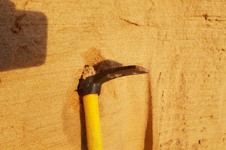 A robust yellow-handled pickaxe digs into a warm, textured sand embankment, suggesting diligent labor. The sun casts a gentle glow across the granular surfaceの写真素材