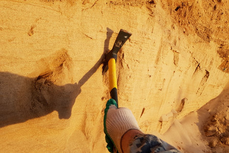 A gloved hand expertly wields a bright pickaxe, carving into sunlit sandy earth. The tool's shadow stretches, revealing diligent excavation effortsの写真素材