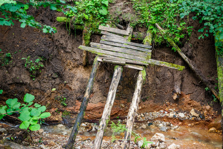 Ramshackle, mossy wooden footbridge precariously spans a tiny creek, leaning against an eroded dirt bank. Old and neglectedの写真素材