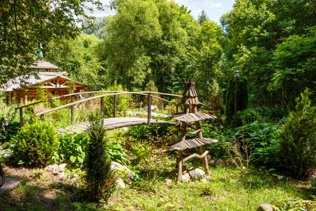 Charming wooden bridge spans over a verdant garden, featuring a quirky rustic wooden art piece. A cabin cozy nestles amidst lush greenery, idyllic summer sceneryの写真素材