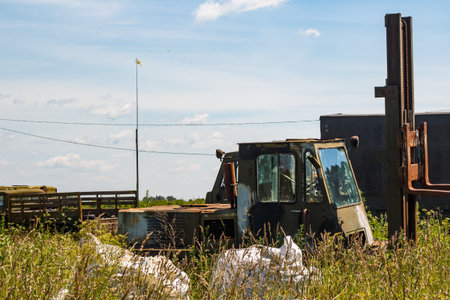 An old, rusty, abandoned forklift truck sits in an overgrown field under a bright sky. This vintage...