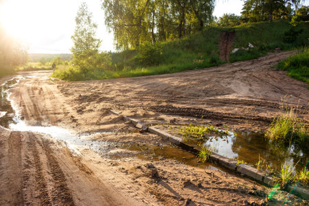 Rugged dirt path with watery ruts and glinting puddles under a bright sun. Tire tracks crisscross unpaved road branching towards green hills, distant treesの写真素材