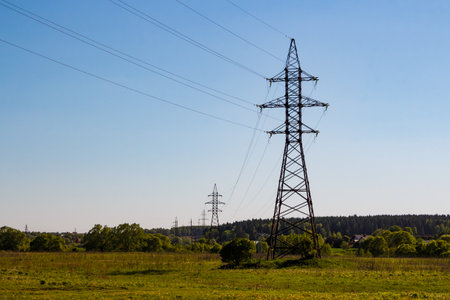 Imposing metal power line tower stands tall in a lush green field. Its robust structure supports numerous high-voltage lines against a clear skyの写真素材