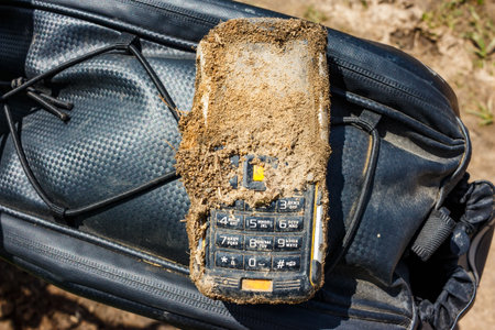 Rugged, mud-caked cell phone sits on a dark textured bag. This tough device endured a messy outdoor...