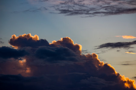 Dramatic dark clouds with glowing golden edges catching vibrant sunset light, contrasting against a soft blue skyの写真素材