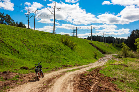 Green railway embankment under a bright sky, with power lines overhead. A solitary bicycle rests on a muddy dirt road, ready for adventureの写真素材