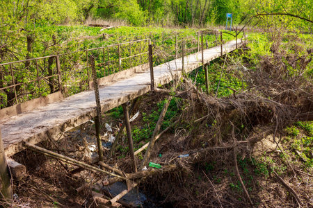 Crooked footbridge over a ditch with a stream, plastic trash in the ditchの写真素材