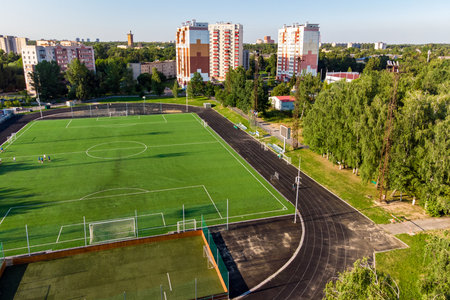 Aerial shot of a well-maintained soccer pitch with a running track and small training ground, flanked by residential high-rises and abundant green trees. An active urban sports areaの写真素材