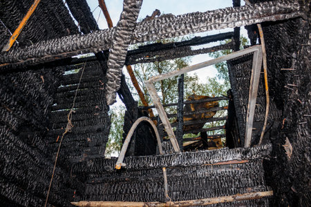 Charred interior of a burnt wooden house, showing blackened beams and walls with glimpses of sky and trees through damaged structureの写真素材