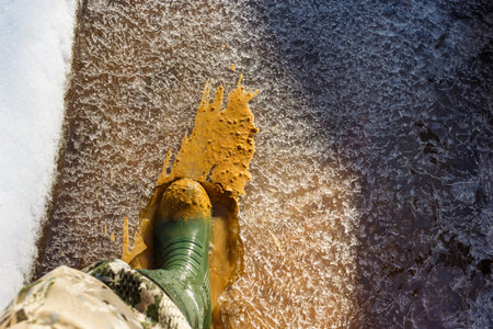 Green rubber boot splashing through a partially frozen stream, breaking ice and splashing muddy slush. Early spring explorationの写真素材