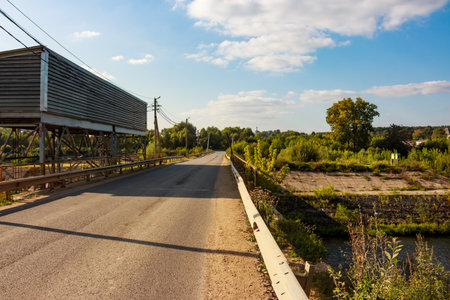 Roadway over a river, featuring waterworks and lush summer greenery under a bright sky. A tranquil rural landscape on a warm dayの写真素材