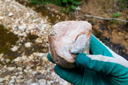 Close-up of a gloved hand clutching a textured flint concretion against a blurred natural stream bed...