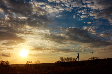 Vibrant sunset sky bursting with golden light and dramatic clouds over a tranquil rural field. Silhouetted trees and power lines mark a peaceful spring eveningの写真素材