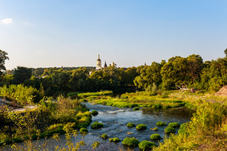 Borovsk monastery's golden domes rise above lush riverbanks and a winding stream under a clear sky,...