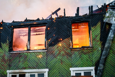 Inferno consumes an old wooden house, fiery glow visible through windows, charred remains of the roof against a smoky sky. Devastating blaze of a derelict buildingの写真素材