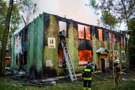 Brave firefighters tackling a raging blaze in an old, abandoned wooden house. Smoke billows from charred windows and a ladder assists in the emergency efforts. Russiaの写真素材