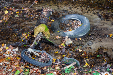 An unsightly mess of discarded tires, a boot, and other rubbish litters a shallow stream, highlighting environmental pollution amid autumn leavesの写真素材