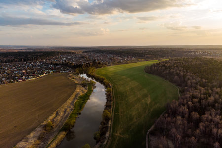 Aerial view of a winding river flowing through vibrant green and brown fields, bordered by a quaint village and dense forest. Tranquil spring evening lightの写真素材