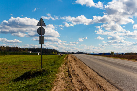 Back of a road sign on a country roadside, flanked by a lush green field and an open asphalt road under a bright blue sky with fluffy cumulus cloudsの写真素材