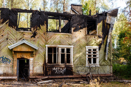 A stark image of a dilapidated, fire-ravaged wooden house. Charred remnants and broken windows evoke abandonment and ruin, standing against a fading autumn backdropの写真素材