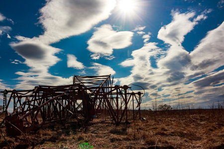 Abandoned, twisted metal remains of a fallen power line tower lie in a dry field, under a dynamic sky with bright sun and cloudsの写真素材