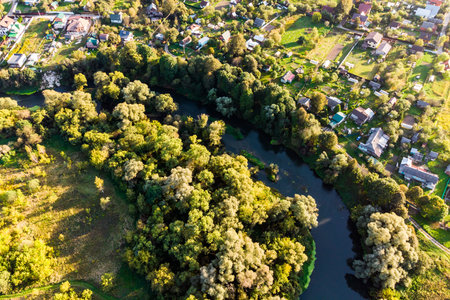 Aerial view of a winding river snaking through lush green trees and a quaint village. Golden hour sunlight bathes the peaceful countryside sceneの写真素材
