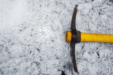 Rustic pickaxe with bright yellow handle rests on a frosty expanse of intricate ice crystals and fresh snow. A scene of winter resilience or cold endeavorの写真素材