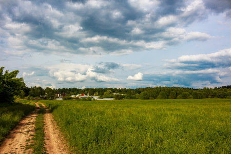 A rustic dirt path meanders through vibrant green fields under a dynamic cloudy sky. This serene rural landscape beckons exploration on a bright summer dayの写真素材