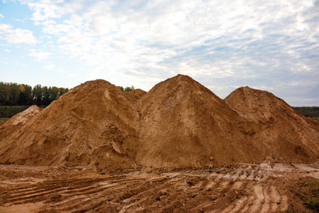 Huge earthen mounds dominate a quarry site under a cloudy autumn sky, showing fresh tire tracks on the muddy ground, depicting aggregate raw material storageの写真素材
