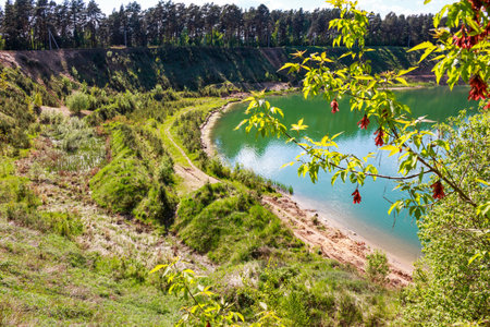 Emerald water fills a stunning quarry lake, embraced by lush green slopes under a sunny sky. A hidden natural gem offering tranquil beauty and a winding path to exploreの写真素材