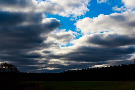 Moody, brooding clouds blanket a vivid blue sky. Striking light breaks through, illuminating sections above a stark, silhouetted forest and a quiet, open fieldの写真素材