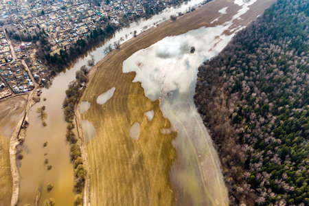Aerial view of flooded spring farmlands next to a swollen river and a dark forest edge near a villageの写真素材